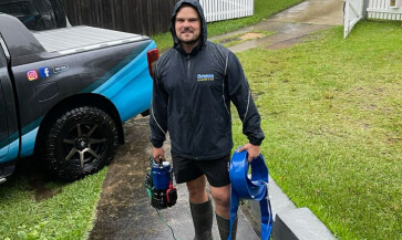 Ben Soutter the owner of dream in a raincoat and gum boots holding a pump, standing infront of the dream plumbing ute ready to tackle a plumbing emergency